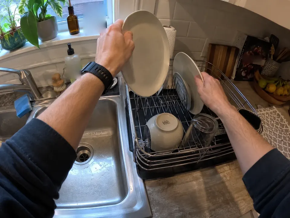 First-person view of hands loading plates into a dish rack beside a kitchen sink.