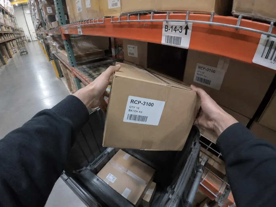 First-person view of hands lifting a labelled carton from a warehouse shelf into a tote.